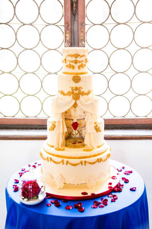 Close-up detail shot of a five-tier wedding cake displayed on a round table covered in a royal blue linen. The cake is ivory with elaborate gold fondant baroque-style scrollwork, swags, and ornamental details on every tier. The center tier features a painted fondant panel depicting an interior ballroom scene with a red rose under a glass dome, framed by ivory fondant curtains draped with gold trim. The cake sits on a red base board. Scattered across the table are deep red rose petals, and a small glass cloche containing a red rose is placed beside the cake. The background features a decorative circular iron window grille, suggesting an indoor venue with architectural detailing. The overall design theme is clearly inspired by the Beauty and the Beast fairytale, with a classic, ornate French baroque aesthetic.