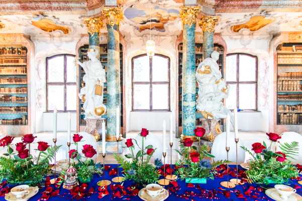 A styled wedding reception tablescape photographed inside an ornate Baroque library or palace hall featuring floor-to-ceiling bookshelves, gilded stucco ceiling details, tall green marble columns with gold capitals, and white classical statues. The long dining table is dressed with a deep royal blue tablecloth and decorated with red roses, scattered rose petals, fern greenery, and tall thin gold taper candlesticks with white candles. Place settings include white teacups and saucers with gold-rimmed charger plates, and small decorative gold and red figurines are placed along the table runner. The overall decor palette is bold red, royal blue, and gold with a dramatic, fairy-tale-inspired theme. Wide shot taken from a slightly elevated angle facing the table head-on. Potential venue feature image.