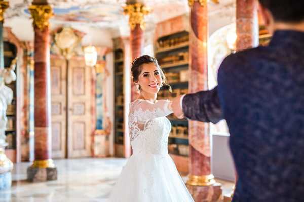 A couple shares a dance or twirl inside an ornate baroque-style library or palace hall, with towering pink marble columns with gilded capitals, painted frescoed walls, and dark wood bookshelves visible in the background. The bride wears a white ball gown with a sheer lace long-sleeved bodice and an illusion neckline, her dark hair styled up with drop earrings, and she is smiling toward the camera. The groom, visible from behind in the foreground wearing a dark navy patterned jacket, holds her hand as she turns. This is a medium portrait shot with a shallow depth of field, keeping the bride in sharp focus while the groom and background are softly blurred. Potential venue feature image.
