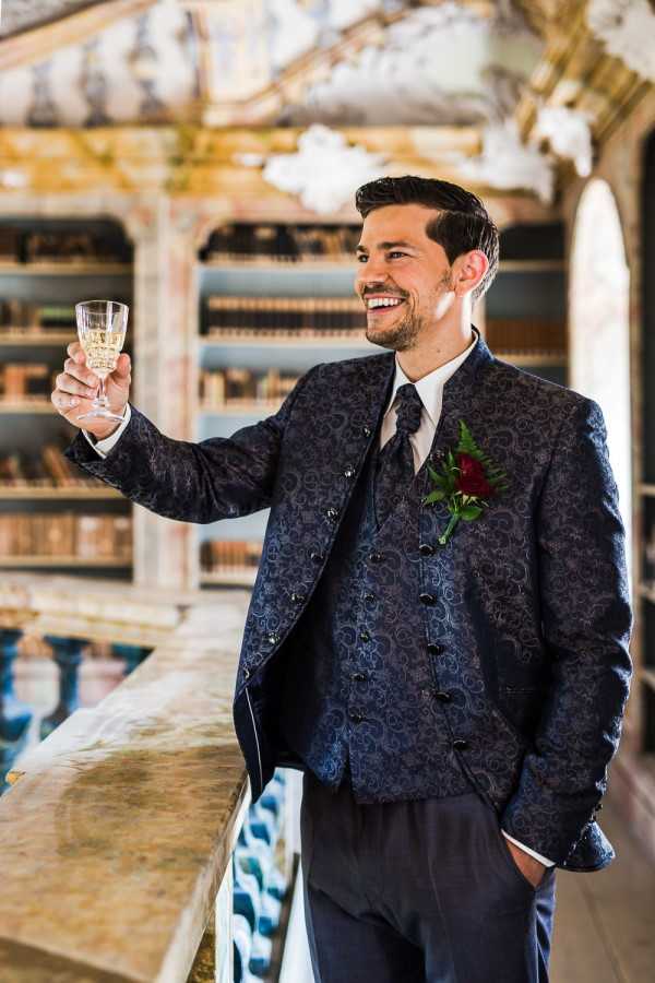 A portrait of a groom raising a crystal champagne flute and smiling, photographed indoors in what appears to be an ornate historic library or reading room with book-lined shelves and decorative frescoed ceiling visible in the background. The groom wears a navy blue paisley brocade jacket and matching waistcoat with black button closures, dark trousers, a white dress shirt, and a dark tie. His boutonniere features a deep crimson red rose with fern greenery. The shot is a medium portrait with soft background blur keeping focus on the groom.