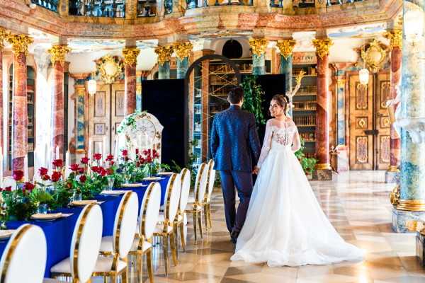 A bride and groom stand hand-in-hand inside an ornate baroque-style library or palace hall, viewed from behind as they look toward the elaborately decorated interior featuring pink and green marble columns, gilded detailing, and floor-to-ceiling bookshelves. The bride wears a white off-the-shoulder ball gown with a long train, and the groom is dressed in a dark navy patterned suit. A long reception table runs along the left side of the frame, draped in deep royal blue linen and set with gold charger plates, tall candles, and a lush runner of red roses and dark green foliage; gold oval-back chairs line both sides of the table. The composition is a wide portrait shot capturing both the couple and the full grandeur of the venue interior. Potential venue feature image.