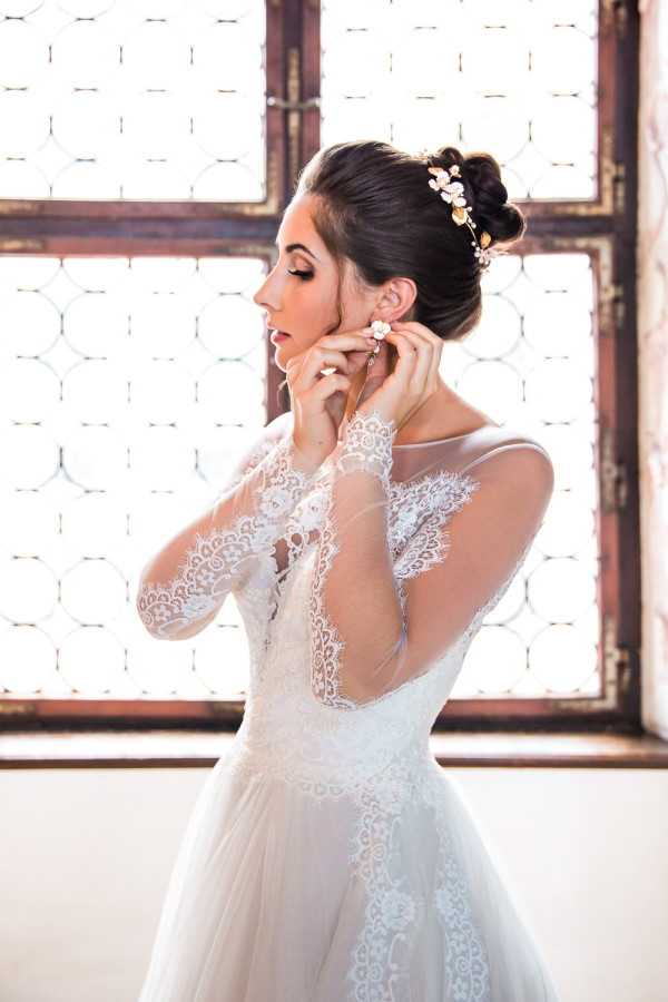 A bride is photographed getting ready, adjusting a small floral drop earring at her ear in a close-to-medium portrait shot. She wears a white long-sleeved wedding gown with sheer illusion lace sleeves and an open lace back panel, paired with a full tulle skirt. Her dark hair is styled in an updo adorned with a gold and blush floral hair piece. She is positioned in profile against a large ornate latticed window that provides bright backlight, set within a rustic interior with aged wooden window frames. The styling is classic with romantic lace detailing.