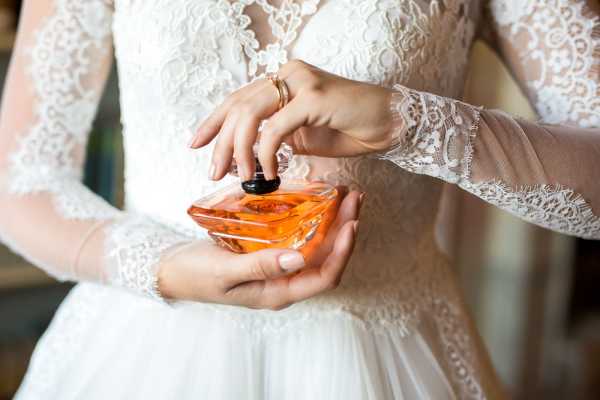 A close-up detail shot of a bride getting ready, showing her hands holding an amber-colored glass perfume bottle with a black cap while a second person's hand assists. The bride is wearing a white long-sleeve lace gown with intricate floral lace detailing on the sleeves and bodice, and gold rings are visible on her fingers. The composition is tightly framed on the hands and midsection, with a softly blurred indoor background.