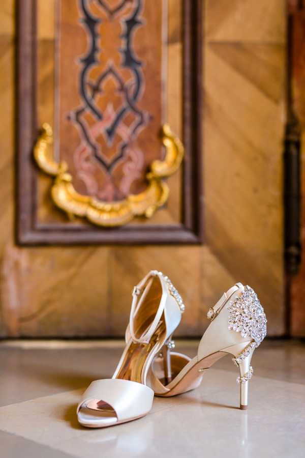 Close-up detail shot of a pair of ivory satin bridal heels with crystal-embellished heel and back strap detailing, placed on a light marble or stone surface. The shoes feature an open toe, slim stiletto heel, and a large rhinestone brooch-style ornament at the back. The background shows an ornate wooden door panel with gold gilt decorative hardware in a baroque or classical style, rendered slightly out of focus to keep the shoes as the primary subject.
