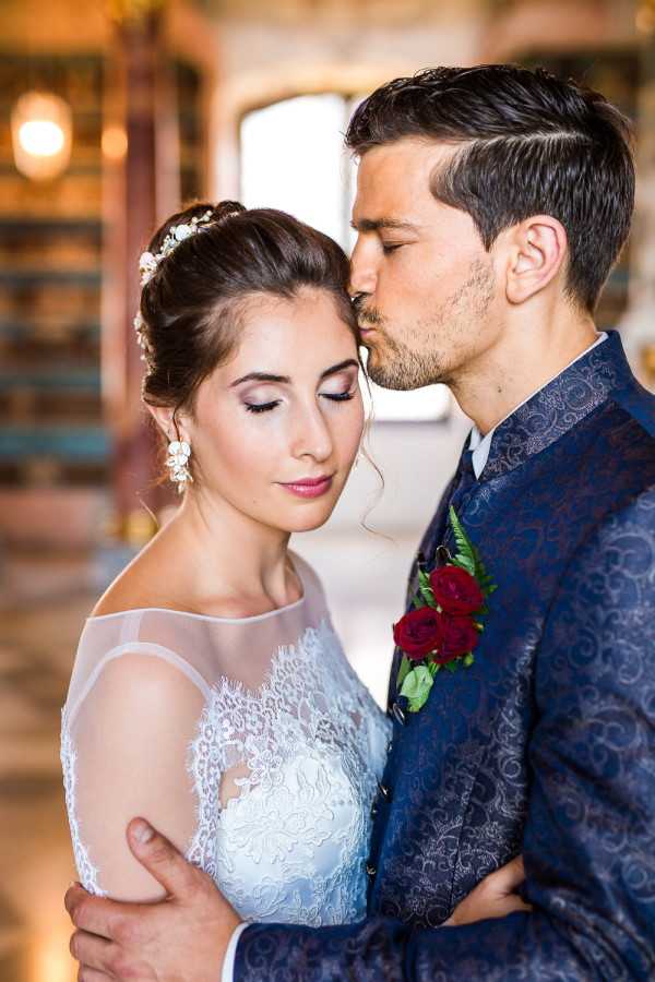 A close-up portrait of a bride and groom in an indoor setting with warm, softly blurred architectural details visible in the background. The groom gently kisses the bride's temple while embracing her from behind; both have their eyes closed. The bride wears a white lace wedding dress with sheer off-shoulder sleeves and an updo hairstyle adorned with a delicate floral hair piece and drop floral earrings. The groom wears a navy blue jacquard-patterned suit jacket with a boutonnière featuring deep red roses and green foliage. The overall styling leans toward a classic-romantic aesthetic with a rich, jewel-toned accent color in the florals.