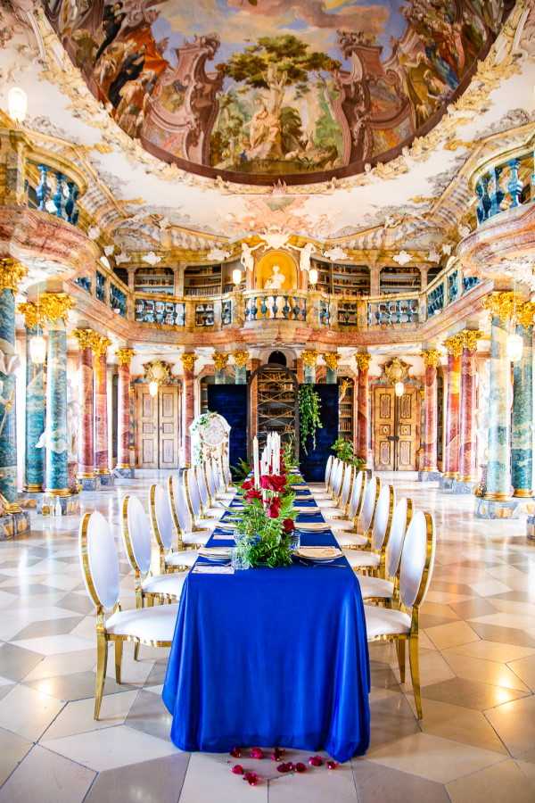 A reception table setup styled inside an ornate Baroque library or palace hall featuring floor-to-ceiling frescoed ceilings, gilded stucco ornamentation, marble columns in deep red and green tones, and two-tiered bookshelves. A long rectangular dining table is dressed in a royal blue satin tablecloth and lined with approximately 16 white cushioned chairs with gold frames. The table runner features lush greenery and deep crimson red roses interspersed with white taper candles. Scattered red rose petals are visible on the marble tile floor at the base of the table. The overall decor palette is royal blue, gold, and deep red, styled in a classic, maximalist theme that complements the room's Baroque architecture. Wide shot taken from the foot of the table looking toward the far end of the hall. Potential venue feature image.