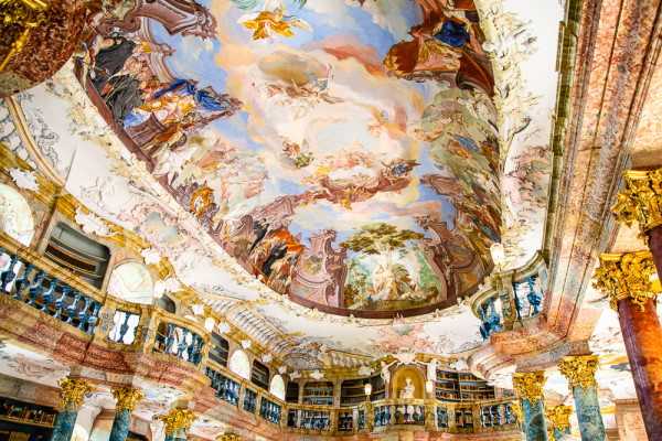 This image shows the interior of a grand Baroque hall, captured from a low upward-looking angle emphasizing the ornate ceiling fresco and architectural details. The ceiling is painted with large-scale figurative scenes featuring angels, clouds, and classical figures in soft blues, pinks, oranges, and golds. The walls feature gilded stucco ornamentation, marbled columns in deep red and green tones, and built-in bookshelves with blue-painted balcony railings across two levels, suggesting this is a historic library or palace hall. No people, couple, or wedding activity are visible in this image. Potential venue feature image.