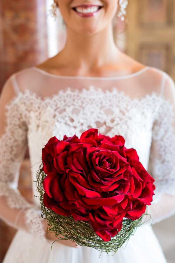 Close-up portrait of a bride holding her bouquet, shot indoors against a warm-toned brick background. The bouquet is a tightly arranged cluster of deep red roses with a collar of curled green wire grass or bear grass wrapping the stems. The bride wears a white long-sleeve lace gown with an illusion neckline featuring delicate floral lace detailing across the bodice and sheer lace sleeves. She is smiling and wearing drop earrings, with red lipstick complementing the bouquet color. The composition is a mid-close-up with the bouquet in sharp focus in the foreground and the bride's face softly focused in the background.
