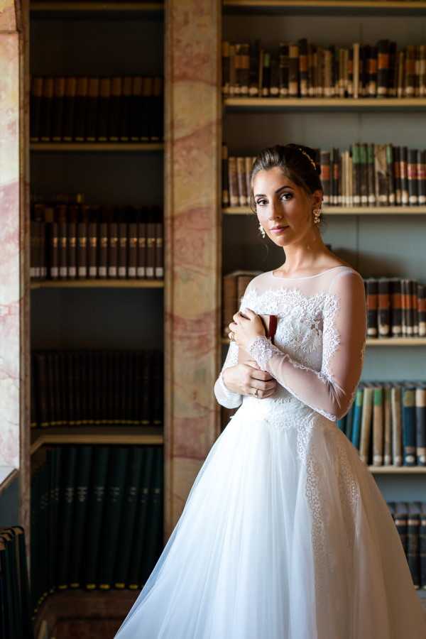 A bridal portrait taken indoors in what appears to be a historic library or study, with floor-to-ceiling wooden bookshelves filled with antique leather-bound books and a marble or stone column visible to the left. The bride stands alone, holding a small dark red object — possibly a book or clutch — close to her chest with both hands. She wears a white ballgown with an illusion neckline, long sheer lace-appliquéd sleeves, and a full tulle skirt. Her dark hair is pulled up with a delicate headpiece, and she accessorizes with chandelier earrings and a ring. The composition is a medium portrait shot with soft natural light coming from the left, creating gentle contrast against the darker bookshelf background.