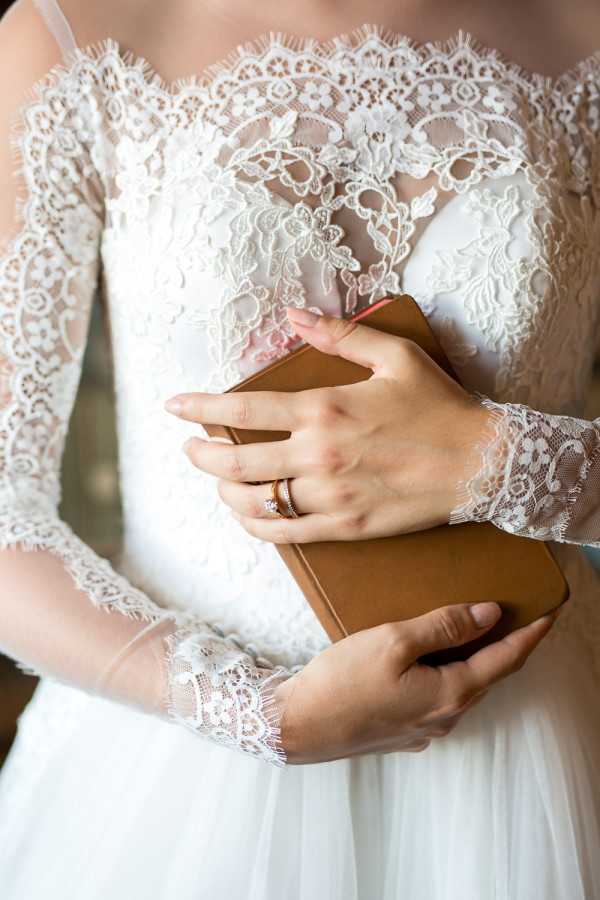 Close-up detail shot of a bride holding a tan leather-bound book, likely containing her wedding vows. She is wearing a white lace gown with an illusion neckline, floral lace appliqué bodice, and long sheer lace sleeves with scalloped cuffs. A rose gold engagement ring and wedding band are visible on her left hand. The composition is tightly cropped to the torso and hands, emphasizing the dress detailing and the rings.