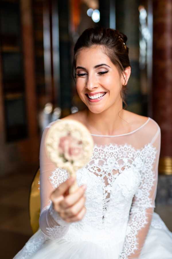 A getting-ready portrait of a bride seated indoors, smiling as she looks down at a small ornate handheld mirror she holds toward the camera. She is wearing a white long-sleeved lace wedding dress with a sheer illusion neckline and intricate floral lace detailing across the bodice. Her dark hair is styled in an updo with soft face-framing pieces, and her makeup includes defined brows, winged eyeliner, and a warm nude lip. The background is softly blurred but suggests an indoor setting with warm wood tones and gold accents. The composition is a close-up portrait with the mirror slightly out of focus in the foreground.