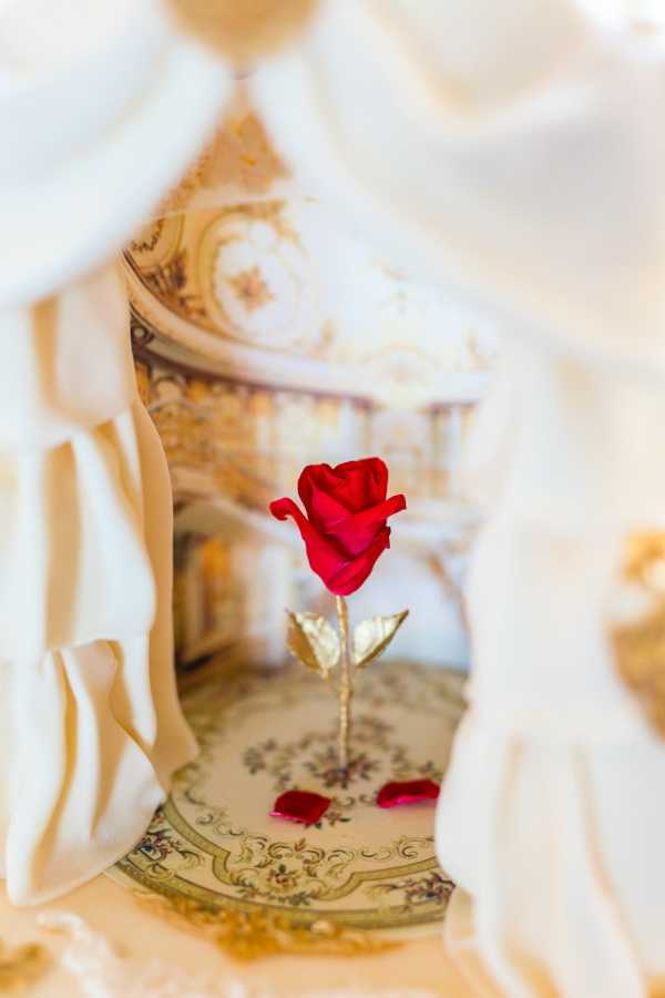 A close-up detail shot of a single artificial red rose with a gold metallic stem and leaves, placed on an ornate cream and gold decorative plate or charger with floral detailing. A few scattered red petals surround the base of the rose. The composition is framed by soft, out-of-focus cream fabric draping in the foreground, and the background shows a miniature or painted baroque-style architectural setting with cream and gold tones, suggesting a fairy-tale or Beauty and the Beast themed styled shoot or wedding decor concept. The overall decor palette is cream, gold, and red, consistent with a classic fairy-tale styling theme.