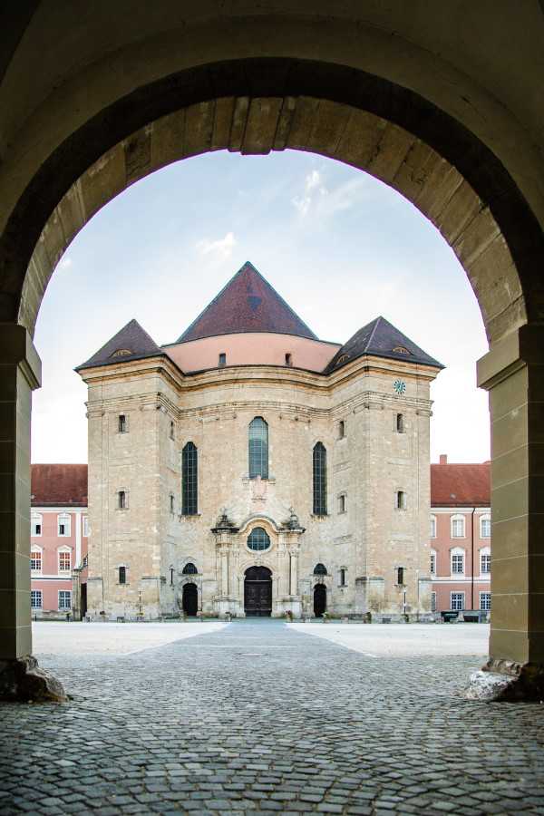A wide-angle architectural shot framed through a large stone archway, looking across a cobblestone courtyard toward a substantial historic church or chapel building. The structure features pale sandstone masonry, tall arched windows, a dark wood ornate baroque entrance portal, a salmon-pink rendered upper section, and multiple steep dark red/brown tiled pyramid-shaped roof towers. Flanking wings of pink-rendered buildings with red tile roofs are visible on either side. No people are present in the image. The composition uses the archway as a natural frame, creating strong symmetry and depth. Potential venue feature image.