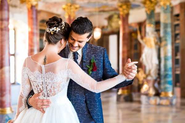 A couple shares a first dance in an ornate palace or château ballroom featuring tall marble columns with gold gilded detailing, painted ceilings, and classical statuary visible in the background. The bride wears a long-sleeve illusion lace gown with a low open back and small button closures, her hair styled in an updo adorned with a gold floral hair piece, and a small tattoo is visible on her upper back. The groom wears a navy blue patterned jacquard suit with a coordinating tie and a deep red rose boutonniere. The composition is a medium portrait shot taken from slightly behind the bride, with the groom smiling warmly as they hold hands in a dance hold.
