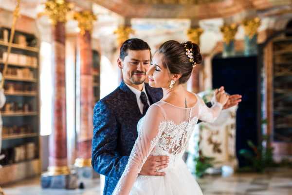 A couple poses together in a portrait shot inside an ornate baroque-style library or palace hall, featuring gilded gold architectural details, tall bookshelves with aged volumes, and pink-hued marble columns. The bride wears a white gown with an illusion lace bodice and sheer long sleeves, her dark hair styled in an updo adorned with small floral pins, and she wears chandelier earrings. The groom is dressed in a deep navy blue jacquard-patterned suit with a dark tie. The composition is a mid-length portrait with the background softly blurred, emphasizing the couple in a classic, formal styling theme. Potential venue feature image.