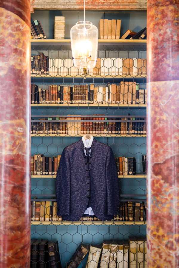 A groom's outfit detail shot taken indoors in what appears to be a library or book-lined room. A navy blue brocade jacket with a subtle floral or paisley jacquard pattern is displayed on a hanger against built-in bookshelves painted teal-blue, filled with aged leather-bound books. A white dress shirt and a gold wooden bow tie are visible beneath the jacket. The shelves are fronted with hexagonal wire mesh, and a glass pendant light with a brass fixture hangs above. Two tall reddish-brown marbled columns frame the composition on either side. The shot is a vertical portrait-style detail image highlighting the groom's attire styling choices within a classic, book-filled interior setting.