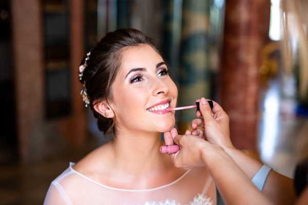 A close-up portrait of a bride during the getting-ready phase, having lip gloss applied by a makeup artist whose hands are visible at the edges of the frame. The bride wears a white lace-trimmed dress with a sheer neckline and has her dark hair styled in an updo adorned with small floral hair accessories. Her makeup includes defined brows, smoky eye shadow, and a soft pink lip. The setting appears to be indoors with warm, diffused lighting and a softly blurred background suggesting a rustic interior space.