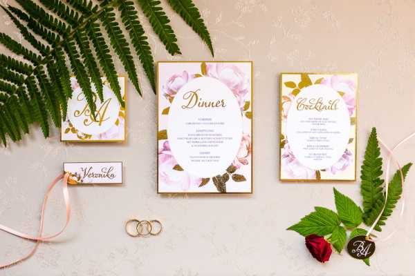 A flat lay composition of a wedding stationery suite arranged on a neutral blush-toned surface. The suite includes a dinner menu card, a cocktails menu card, a monogram card, and a place card reading 'Veronika,' all featuring a consistent design of blush pink roses, olive green foliage, and gold script typography with gold border details. Accompanying the stationery are two gold wedding bands, a coiled blush pink satin ribbon, a fresh dark red rose bud, a green wax seal tag with gold monogram initials, and two fern fronds used as styling props.