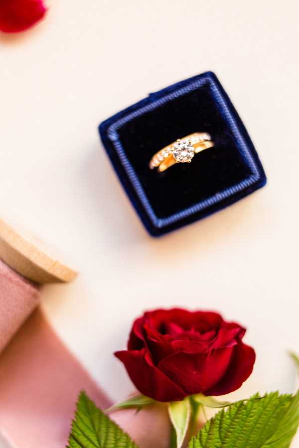 A detail close-up flat lay showing a yellow gold engagement ring with a round solitaire diamond and pavé-set side stones, displayed in an open navy blue velvet ring box against a cream surface. A single deep red rose is positioned in the lower portion of the frame, and the corner of a blush pink gift box is partially visible on the left side. The styling uses a classic red and navy color palette, suggesting a formal or traditional wedding aesthetic.