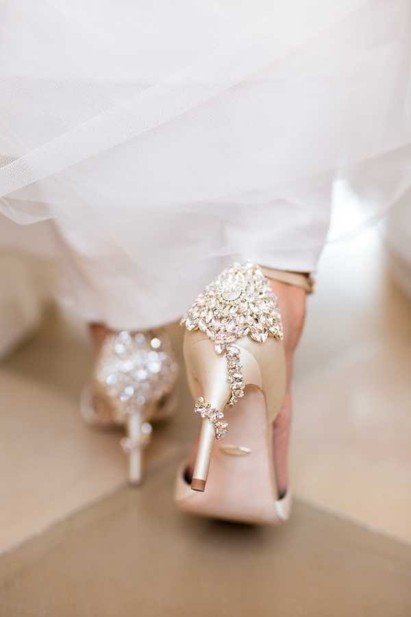 Close-up detail shot of the bride's shoes during a getting-ready moment. The bride is wearing champagne/blush satin heels adorned with large crystal and rhinestone embellishments on the back — a central starburst cluster of marquise-cut and round stones with a trailing vine of smaller crystals extending down toward the heel. The white tulle of the bride's wedding dress and veil drape softly over and around the shoes. The composition is a tight rear-facing detail shot focusing entirely on the decorative heel embellishments, with shallow depth of field blurring the background floor.