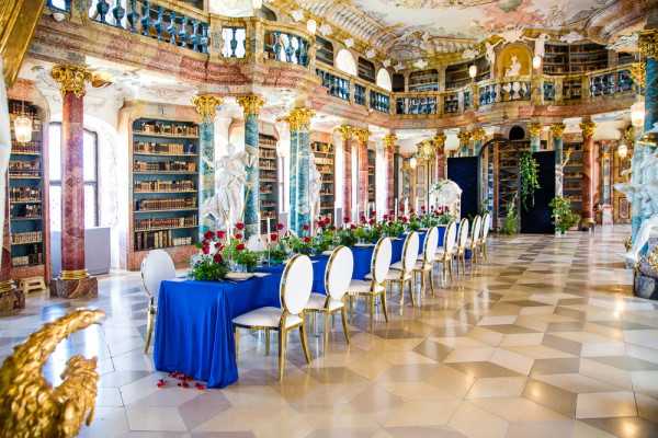 A wedding reception table is set up inside a grand Baroque library hall featuring floor-to-ceiling bookshelves, ornate gold-gilded columns with marbled finishes, painted ceiling frescoes, and classical white marble statues. The long rectangular dining table is draped in a cobalt blue tablecloth and lined with white oval-backed chairs with gold frames on either side. Centerpieces consist of lush arrangements of deep red roses and green foliage running the length of the table, with scattered red rose petals on the polished checkered marble floor. No guests or wedding party are present in this wide-angle shot, which primarily showcases the reception setup within the architectural interior. Potential venue feature image.