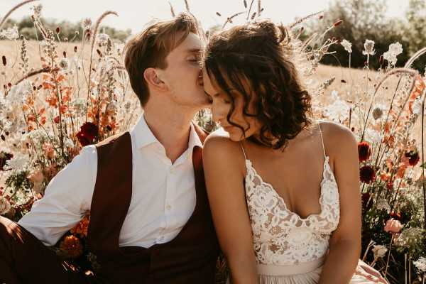 A couple shares a kiss during an outdoor portrait session, seated among tall wildflowers in a field setting. The groom wears a white dress shirt with a dark brown vest, while the bride wears a spaghetti-strap lace bodice wedding dress with her dark curly hair worn half-up. The surrounding wildflowers include deep burgundy and coral-red poppies, blush ranunculus, and white cow parsley, creating a rich warm-toned boho aesthetic. The shot is a close-up portrait with soft warm backlighting, giving the image a golden haze consistent with late afternoon sun.