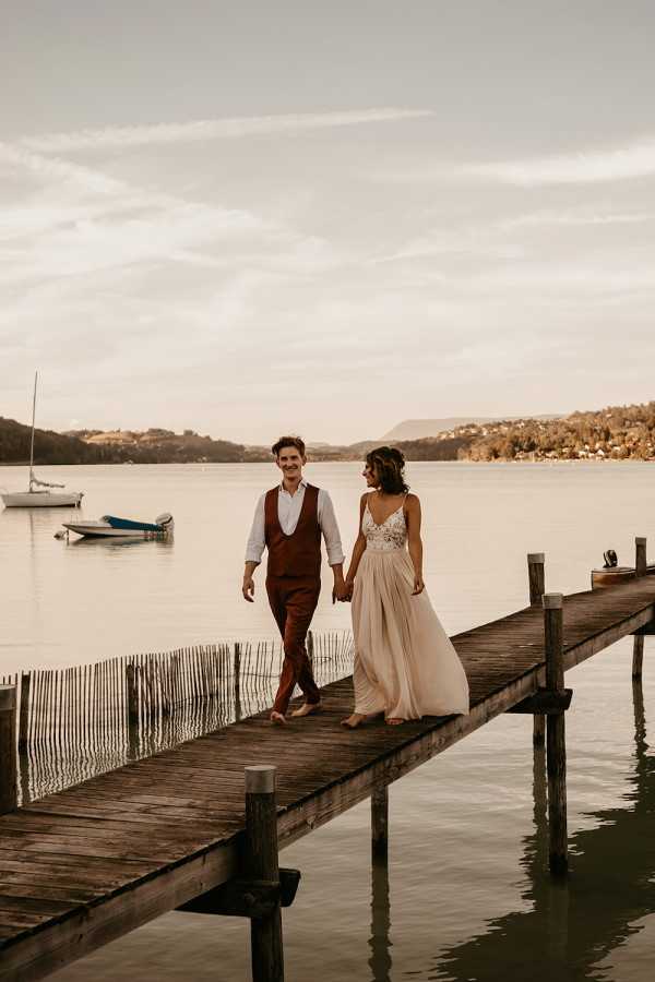 A couple walks hand-in-hand along a wooden dock extending over a calm lake, both barefoot and smiling at each other. The bride wears a flowing champagne-toned gown with a lace bodice and a lightweight, flowy skirt, while the groom is dressed in rust-brown trousers and a matching waistcoat over a white open-collar shirt. The setting is outdoors on a lake with small sailboats visible in the background, giving the portrait a relaxed, boho aesthetic. The shot is a medium-wide portrait framing both figures in full length with the lake and distant hillside as the backdrop.