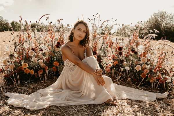 A bridal portrait of a single woman seated on dry hay in an outdoor field setting, with one knee raised and her dress train spread around her. She wears a slip-style wedding dress in ivory/champagne with a lace bodice and thin spaghetti straps. Behind her is a styled floral arrangement at ground level featuring burnt orange dahlias, deep burgundy blooms, terracotta and blush flowers, pampas grass, and dried grasses in warm autumn tones, creating a boho aesthetic. The shot is a medium portrait with warm golden-hour lighting, and the overall styling palette is earthy and autumnal.