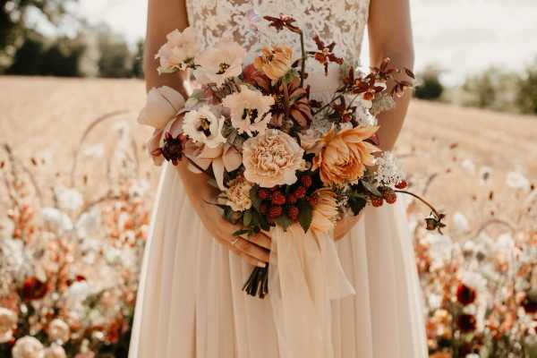 Close-up portrait of a bride from the neck down, holding a bridal bouquet outdoors in a field setting. The bride wears an ivory lace bodice top and a flowing ivory chiffon skirt with a wide ribbon wrist cuff. The bouquet features a rich autumnal palette of peach garden roses, burnt orange dahlias, cream lisianthus, deep burgundy hellebores, rust-toned carnations, and clusters of red berries, with trailing green foliage and long ivory satin ribbons. The arrangement has an unstructured, garden-style composition with branching elements extending outward. The background shows a warm golden wheat field with scattered wildflowers in terracotta, cream, and burgundy tones, consistent with a late-summer or early-autumn shoot.