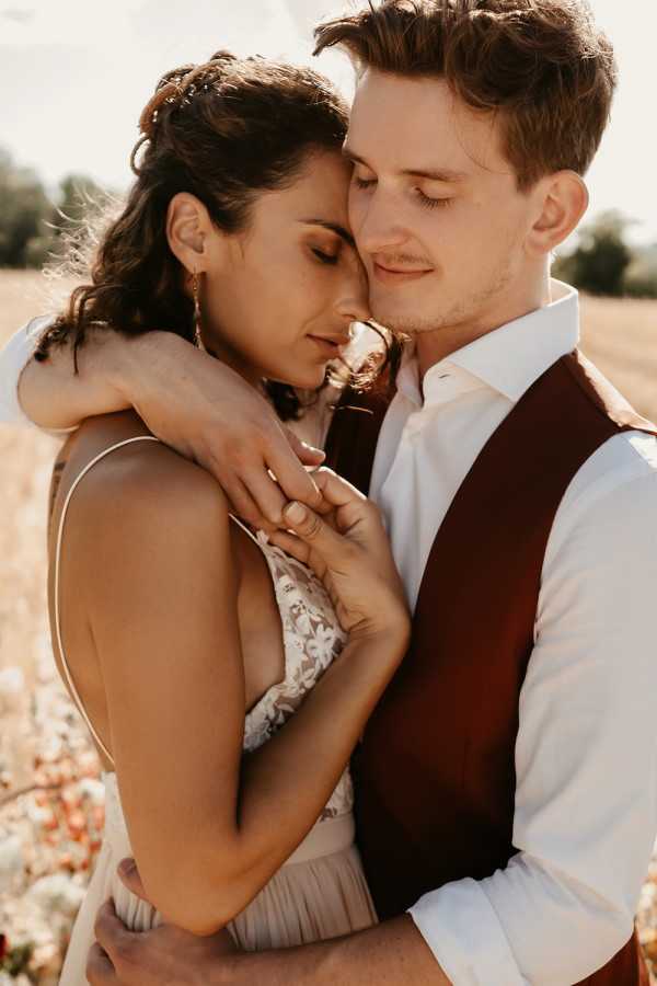 A close-up portrait of a couple embracing outdoors in what appears to be an open field, both with eyes closed in an intimate moment. The bride wears a thin-strap dress with delicate floral lace detailing and has dark wavy hair worn half-up with small hair accessories and gold drop earrings. The groom wears a white dress shirt paired with a deep burgundy waistcoat and has light brown hair with light stubble. The warm golden light suggests late afternoon or golden hour shooting. The styling leans toward a relaxed, romantic boho aesthetic with warm earthy tones — the burgundy waistcoat and neutral lace dress forming a cohesive palette. Shot in a tight portrait crop with soft background bokeh.