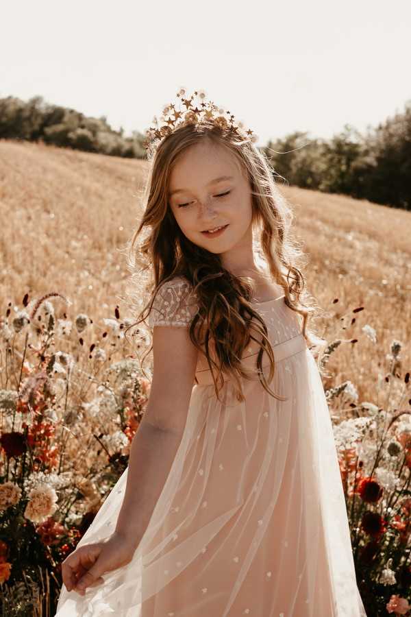 A flower girl is photographed outdoors in a field setting, surrounded by wildflowers including deep red dahlias, white blooms, and coral flowers in the foreground. She is wearing a blush pink tulle dress with cap sleeves and a small star or dot pattern on the fabric, accessorized with a gold star-shaped crown headpiece. Her long wavy brown hair falls loosely over her shoulders. The portrait is a medium close-up shot with warm golden-hour lighting, giving the image a sun-drenched, warm-toned feel consistent with a boho or whimsical wedding styling theme.