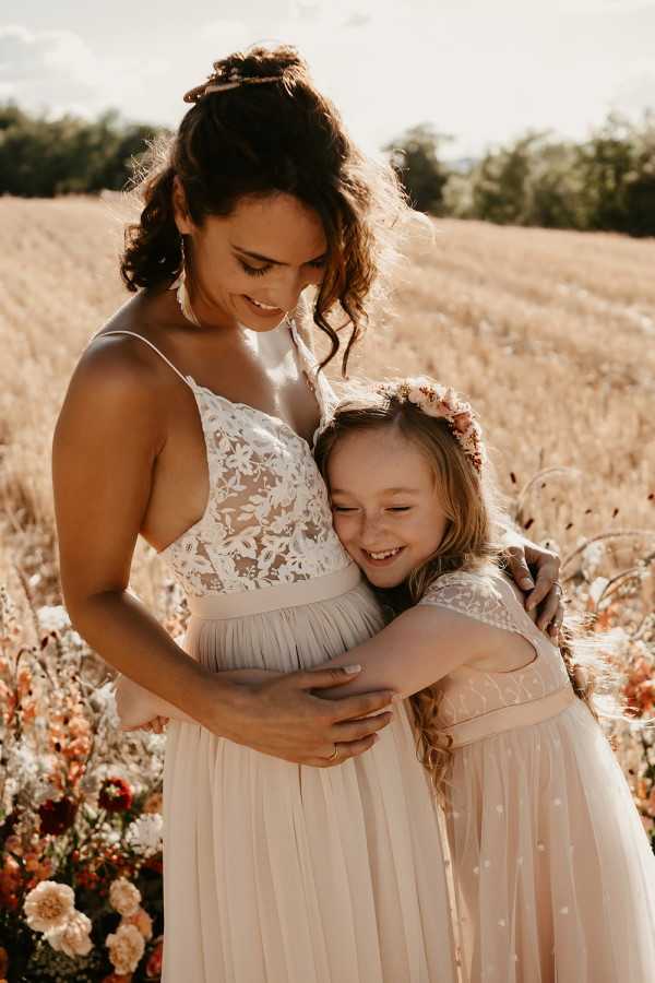 A portrait of a bride and a young flower girl embracing outdoors in a golden wheat field, shot in warm late-afternoon light with a boho styling theme. The bride wears a white floral lace bodice with spaghetti straps over a blush-toned pleated tulle skirt, with her dark hair pinned up in a loose updo. The flower girl wears a blush pink dress with a lace cap-sleeve bodice and a floral crown of terracotta, rust, and blush blooms in her hair. In the lower left foreground, a cluster of florals in peach, rust, and deep burgundy — including what appear to be dahlias and ranunculus — adds color to the composition. The shot is a close-to-mid portrait with warm golden tones.