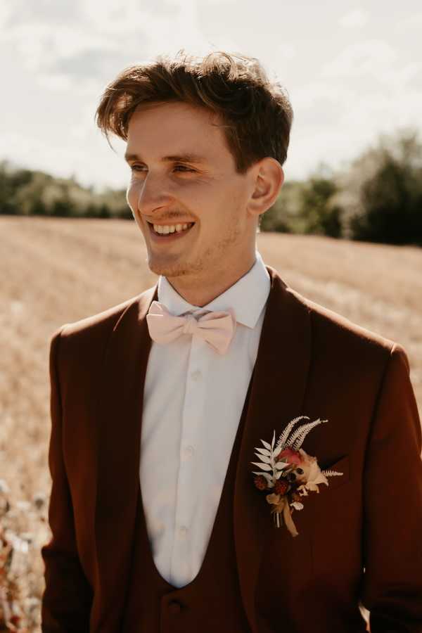A close-up portrait of a groom standing outdoors in a harvested field, smiling and looking slightly off-camera. He is wearing a rust brown three-piece suit with a white dress shirt and a blush pink bow tie. His boutonniere features dried fern fronds, deep burgundy berries, and mixed dried florals in warm terracotta and cream tones, consistent with a boho or earthy autumnal styling theme. The shot is framed from roughly the chest up, with natural golden light creating warm tones across the image.