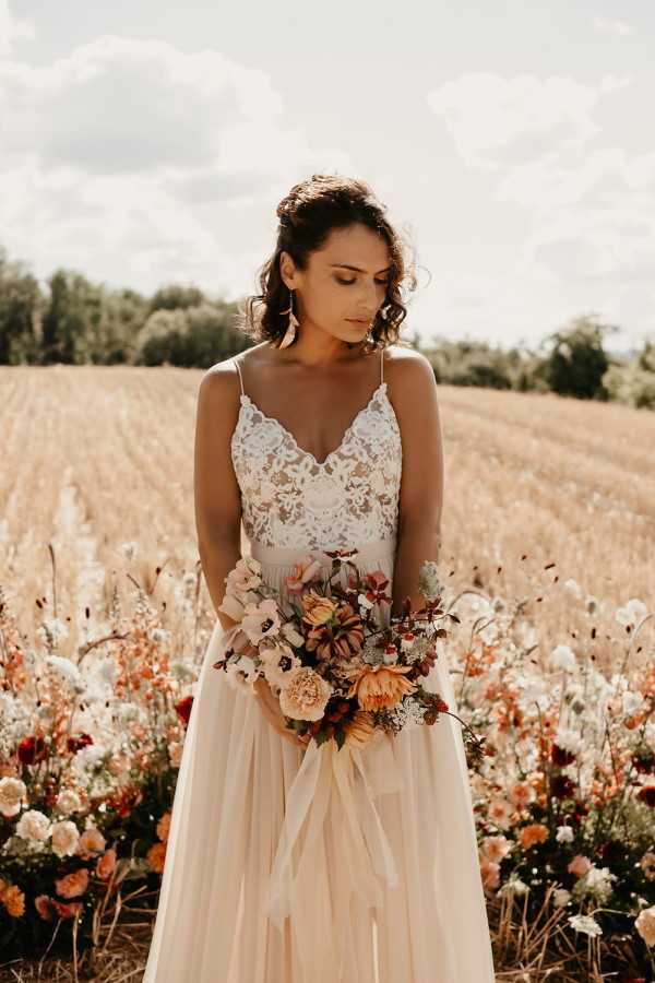 A bridal portrait taken outdoors in a harvested field setting, with the bride standing among a cluster of loosely arranged flowers at ground level. The bride wears a two-piece style gown with a white lace spaghetti-strap bodice featuring floral lace detailing and a flowing champagne/blush skirt. Her dark hair is worn half-up with loose curls, and she looks downward with a relaxed expression. She holds a lush, loosely arranged bouquet featuring burnt orange, terracotta, blush pink, and deep burgundy blooms including what appear to be proteas, garden roses, dahlias, and delicate white filler flowers, tied with a trailing ribbon. The surrounding floral arrangement at ground level echoes the same warm autumn palette of rust, peach, burgundy, and white. The overall styling is boho with a warm autumnal color palette. Medium portrait shot with a shallow depth of field.
