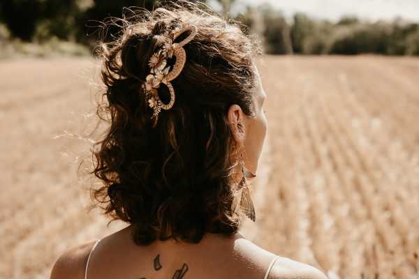 Close-up portrait shot from behind of a bride with curly dark brown hair styled in a half-up look, adorned with a decorative hair accessory featuring woven rattan or straw detailing and small white daisy flowers, consistent with a boho aesthetic. She wears what appears to be a white or ivory thin-strap dress and has visible black tattoos on her upper back and neck area. The image is taken outdoors in bright, warm sunlight against a blurred background of a harvested wheat or grain field.