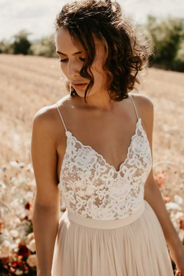 A close-up portrait of a bride standing outdoors in what appears to be a golden wheat field, shot in warm natural light. She wears a two-piece style wedding dress featuring a sheer lace bodice with floral appliqué detailing and thin spaghetti straps, paired with a soft blush-toned pleated skirt. Her dark brown hair is styled in loose curls and pulled partially back, and she is looking downward with a relaxed expression. The edge of a floral arrangement with peach, rust, and cream tones is partially visible at the lower right of the frame, suggesting a boho or romantic outdoor styling aesthetic.