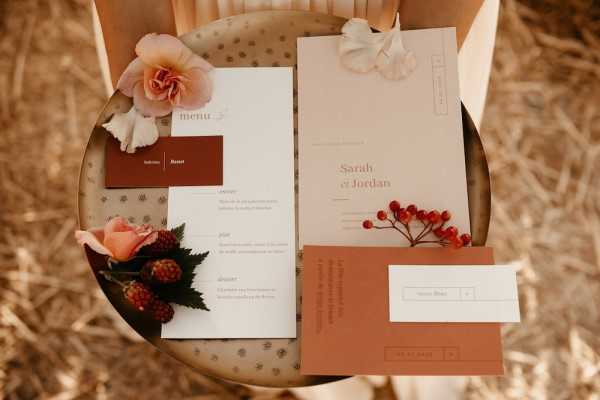 A flat lay detail shot of a wedding stationery suite displayed on a small round decorative table, set against a background of dry golden straw. The suite belongs to a couple named Sarah et Jordan and includes a wedding invitation on warm beige card stock with terracotta typography, a menu card in white with a terracotta accent panel, a terracotta-colored RSVP card, a place card, and a small dark burgundy card. The stationery is styled with a peach garden rose, a white ranunculus or similar ruffled bloom, a small cluster of coral-pink roses with blackberries and green foliage, and a sprig of red berries, all reinforcing an autumn-toned color palette of terracotta, warm beige, and deep rust. The overall styling theme is modern with warm autumnal tones.