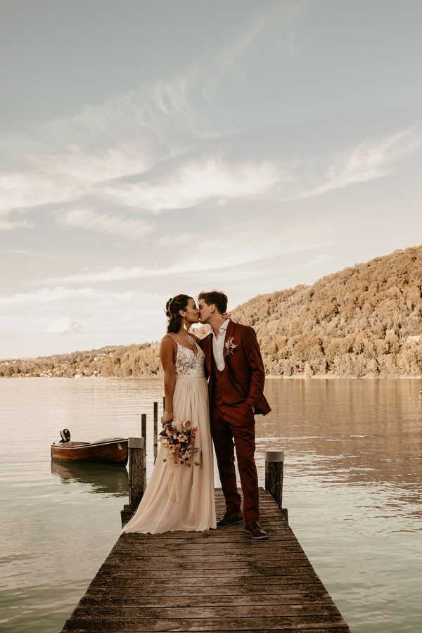A couple shares a kiss during a portrait session on a wooden dock extending over a calm lake, with a small wooden rowboat moored to the left. The bride wears a floor-length gown with a floral embroidered bodice and a soft blush chiffon skirt, holding a bouquet of terracotta, rust, and mauve toned flowers. The groom wears a deep burgundy three-piece suit with a light tie and a matching floral boutonnière. The image is a full-length wide shot with the couple centered on the dock, the lake and tree-covered hillside forming the backdrop, and the photo has a warm, golden-toned edit giving it an autumnal feel.