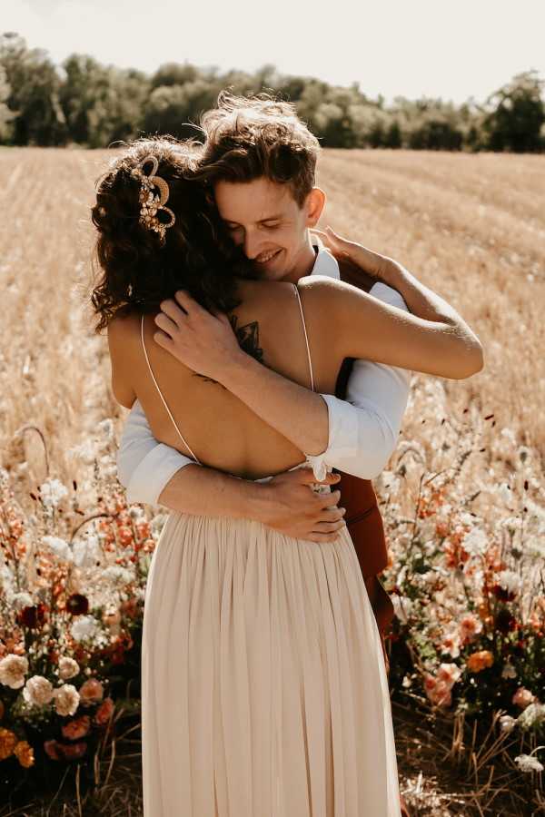 A couple embraces in an outdoor portrait taken in an open wheat field, with clusters of terracotta, rust, ivory, and amber blooms — likely dahlias and ranunculus — arranged at ground level around them. The bride wears a backless, spaghetti-strap champagne/blush gown with a low open back, and her dark curly hair is pinned up with a gold ornamental hair accessory; a small tattoo is visible on her upper back. The groom wears a white dress shirt and what appears to be a rust-brown or tan suit, and is smiling warmly while holding her close. The overall styling has a boho, earthy aesthetic with a warm autumn color palette, and the shot is a medium portrait framed from roughly the waist up.