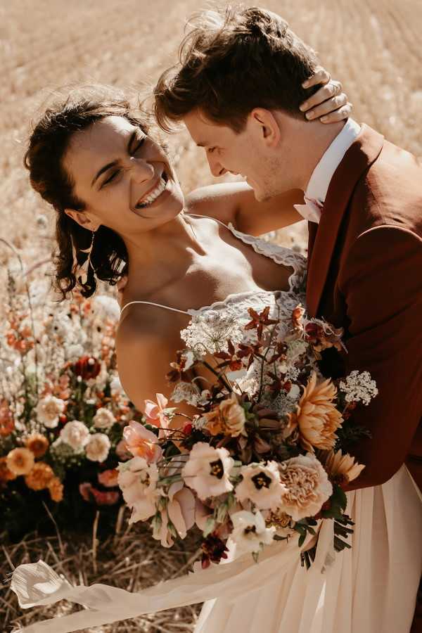 A couple portrait taken outdoors in a dry field setting, with the bride and groom facing each other closely and laughing. The bride wears a spaghetti-strap ivory lace bodice gown and holds a large bouquet featuring burnt orange dahlias, blush garden roses, burgundy tulips, white queen anne's lace, and amber chrysanthemums. The groom wears a rust/terracotta brown suit jacket with a white shirt and a bow tie in a matching warm tone. The overall styling follows a warm autumnal boho palette with earthy rust, blush, and amber tones throughout the florals and clothing. The shot is a close-up portrait with a shallow depth of field, slightly blurred wildflowers visible in the background.