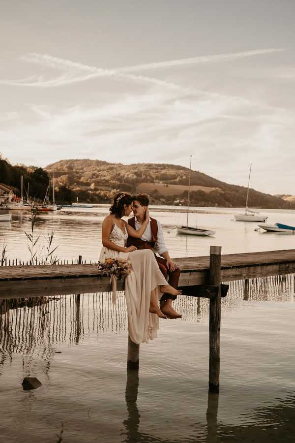A couple sits closely together on the edge of a weathered wooden dock over calm lake water, foreheads nearly touching in an intimate portrait. The bride wears a flowing ivory lace and chiffon dress with a floral hair accessory and holds a loose boho-style bouquet featuring warm terracotta, mustard yellow, and dried tones. The groom is dressed in a white shirt, brown leather vest, brown belt, and brown boots, giving the pair a coordinated rustic-boho aesthetic. The wide-angle portrait is shot at golden hour with sailboats and a hillside visible in the background, warm amber tones washing over the entire scene.