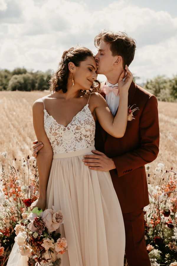 A couple portrait taken outdoors in a field setting, with the groom kissing the bride on the cheek while she smiles. The bride wears a spaghetti-strap wedding dress with a white lace bodice and a flowing champagne-toned skirt, paired with gold drop earrings and an upswept wavy hairstyle. She holds a loosely arranged bouquet featuring blush, dusty rose, burgundy, and peach blooms including garden roses and ranunculus. The groom wears a rust-brown suit jacket with a light pink bow tie and a floral buttonhole. The couple is surrounded by wildflowers in shades of red, coral, and cream. The overall styling is bohemian with warm autumnal tones. Medium portrait composition shot in natural daylight.