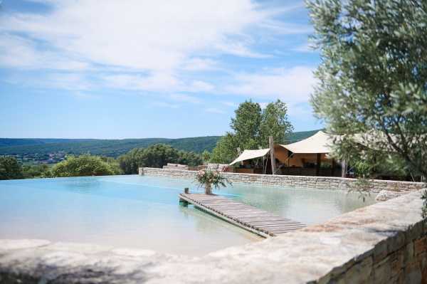 Wide shot of an outdoor infinity pool area with a wooden walkway extending across the water toward a terrace in the background. A cream-colored shade sail canopy is visible over what appears to be a lounge or event area, with wooden furniture underneath. The pool is bordered by stone walls, and an olive tree frames the right side of the image. No people are visible in the shot. Potential venue feature image.
