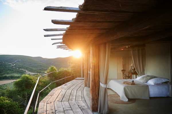 This image does not appear to be a wedding photograph. It shows an open-air accommodation suite or lodge room with a four-poster bed dressed in white and ivory linens, set on a wooden deck with rustic log beam roofing and sheer linen drape panels. The structure overlooks a wide valley landscape at sunset, with warm golden backlight flooding through the beams. The styling is earthy and organic, featuring raw timber construction, a woven jute rug, and neutral tones throughout. Potential venue feature image.