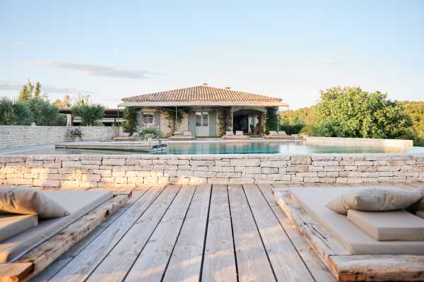 Wide-angle shot of a Provençal stone villa viewed from a wooden deck terrace featuring weathered timber sun loungers with neutral linen cushions in the foreground. A rectangular pool with calm water sits between the deck and the main building, which has a terracotta tile roof, stone facade, and covered loggia. The overall palette is warm sand, pale stone, and soft grey-beige. No people are visible in the image. Potential venue feature image.