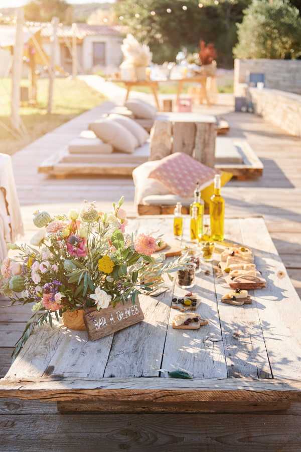 An outdoor cocktail hour or reception food station styled on a rustic whitewashed wooden farm table, featuring a wildflower arrangement in a wooden crate with coral dahlias, pink anemones, yellow blooms, white flowers, and olive branch foliage. A small handwritten wooden sign reads 'Bar à Olives,' and the table is set with boards of bread, olives, and other appetizers alongside bottles of olive oil. In the background, a boho-rustic lounge area is visible with low wooden platform seating, cream and blush scatter cushions, and string lights strung overhead, with a low stone building and additional styled tables further back. The overall styling palette is warm earth tones, blush, and cream with a relaxed, boho-rustic aesthetic. Wide shot taken at golden hour with warm backlit natural light.