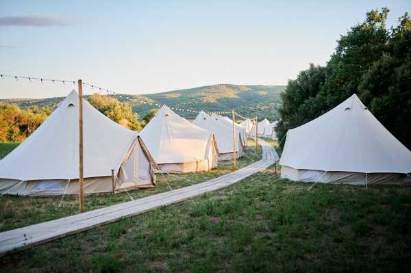 A wide shot of a glamping accommodation setup on an outdoor hillside property, featuring a row of at least eight white canvas bell tents with natural wooden pole supports arranged along a wooden boardwalk pathway. Strings of festoon lights are strung between wooden posts running the length of the tent row. The tents are identical in style, cream-colored with tan canvas lower panels, suggesting a coordinated on-site guest accommodation arrangement for a wedding or multi-day event. The image is taken at golden hour, with warm low light illuminating the scene. Potential venue feature image.
