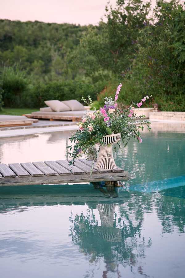 A floral arrangement detail shot at an outdoor pool setting, captured at dusk. A white slatted urn vase holding a loose, garden-style arrangement of pink sweet peas, mauve stock flowers, white blooms, and trailing eucalyptus branches sits on a weathered wooden dock at the pool's edge. The arrangement's reflection is visible in the still turquoise pool water below. In the background, two low-profile wooden sun loungers with beige cushions are arranged on the pool deck. The styling is relaxed and natural, consistent with a rustic or boho aesthetic. Potential venue feature image.