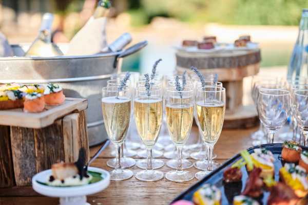 Close-up detail shot of an outdoor cocktail hour spread set on a rustic wooden table. Multiple champagne flutes filled with sparkling wine are arranged in rows, each garnished with a small sprig of dried lavender. Surrounding the glasses are assorted canapés and bite-sized hors d'oeuvres displayed on wooden boards, a tiered wooden cake stand, and small white pedestals, alongside empty wine glasses and a galvanized metal ice bucket with bottles chilling in the background. The styling has a rustic Provençal theme, with the lavender garnish and raw wood serving pieces as intentional design choices.