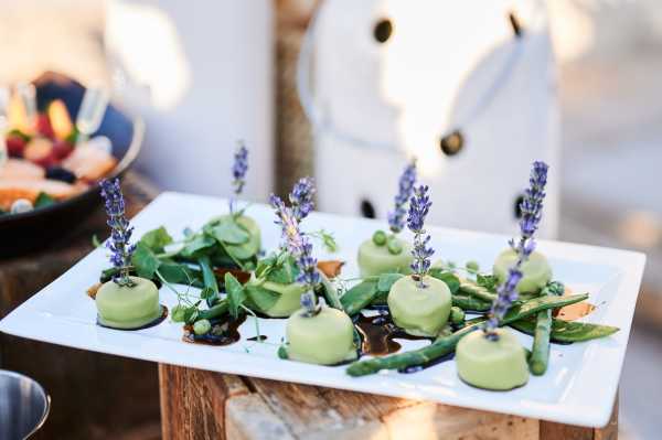 Close-up detail shot of a wedding canapé platter presented on a white rectangular serving plate at what appears to be an outdoor cocktail hour. The platter features several round green-glazed bite-sized portions garnished with fresh lavender sprigs, microgreens, snap peas, and a dark balsamic-style sauce. The food styling uses a green and purple color palette with the lavender as a decorative herb element. The background is softly blurred, revealing a rustic wooden surface and what appears to be additional food platters to the left.